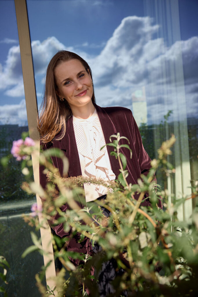 Smiling woman in a burgundy blazer posing outdoors beside a glass wall, with plants in the foreground and a blue sky with clouds behind.