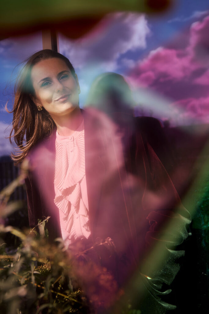Portrait of a woman in a pink blouse and dark blazer, standing outdoors with vibrant rainbow light leaks across the scene.